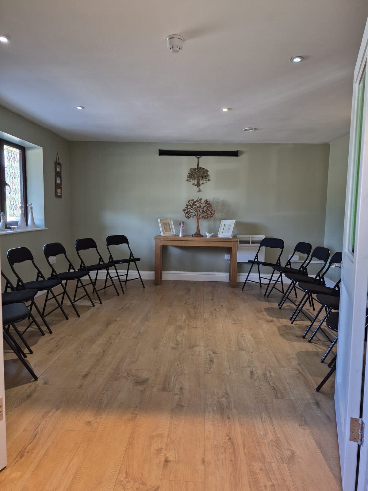A quiet room with black chairs set out in a circle and a small table with two framed photographs and a wooden tree